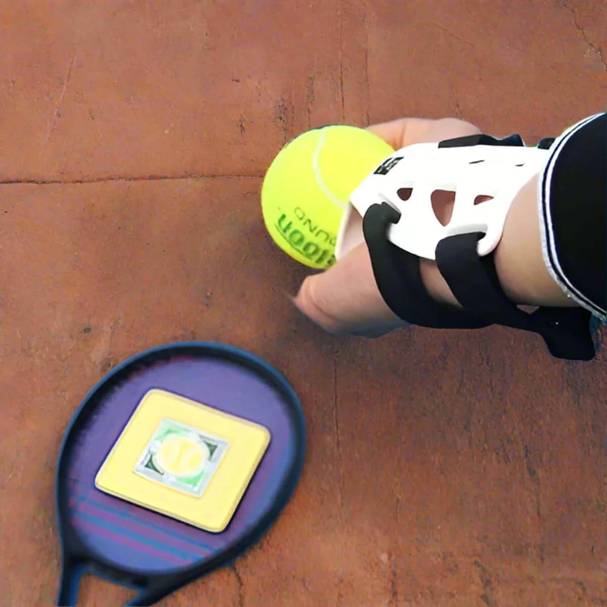Wrist guard support for tennis holding a yellow tennis ball with a racket nearby on a clay court.